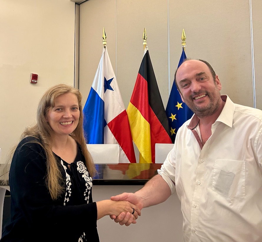 Andrea Meenken (links), Stationsleiterin der Deutschen Seemannsmission Panaman reicht Magnus Deppe die Hand. Deppe leitet die Bremer Seemannsmission. Hinter den beiden sind die Flagge von Panama, die deutsche udn die der Europäischen Union zu sehen. Andrea Meenken trägt ihr langes, blondes Haar offen und ein schwarzes, langes Oberteil. Deppe trägt ein weißes, kurzärmeliges Businesshemd.