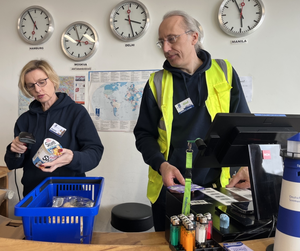 Das Foto zeigt eine Ehrenamtliche und den Leiter der Seafarers' Lounge, Olaf Schröder, an der Kasse der Lounge in Hamburg-Steinwerder. Die Lounge liegt im Kreuufahrtterminal und ist für die Crews der Kreuzfahrtschiffe gedacht. Hinter den beiden an der Wand hängen Uhren mit unterschiedlichen Uhrzeiten und eine Weltkarte. Beide tragen Namensschilder auf ihren dunkelblauen Hoogies. Olaf Schröder trägt auch eine neongelbe Warnweste.