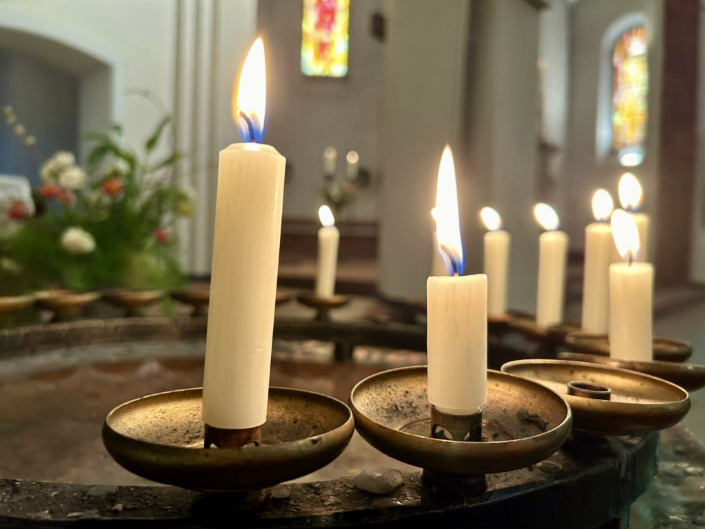 Das Foto zeigt Kerzen, die zum Gebet aufgestellt werden, etwa wenn man in einer Kirchen einem Verstobenen gedenken möchte. Mehrere weiße, schmale Kerzen brennen auf einem Metallständer im Halbkreis.