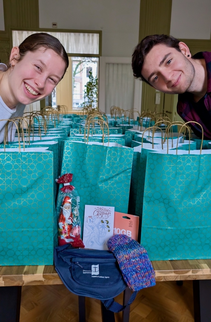 Alma Bloech und Joel Dressler (re.) haben Weihnachtstüten für Seeleute im Amsterdamer Hafen gepackt. Sie lächeln freundlich in die Kamera und haben sich über viele Tüten gebeugt, die auf einem Tisch aufgereiht stehen.