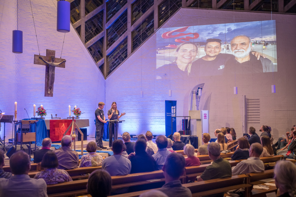 Das Foto zeigt den Innenraum der Auferstehungskirche in Hannover. Im Altarraum stehen Diakon Philipp Manthey und Kathrin Grund von der Seemannsmission in Bremerhaven und Santos. Kathrin Grund spricht gerade ins Mikrofon und stellt den Zuhörern ihre Arbeit in Brasilien vor. Im rechten oberen Rand des Bildes sieht an ein Foto von Kathrin Grund und zwei Kollegen am Strand von Santos. Es ist eine Abendveranstaltung, das Licht ist gedämpft und leicht bläulich.