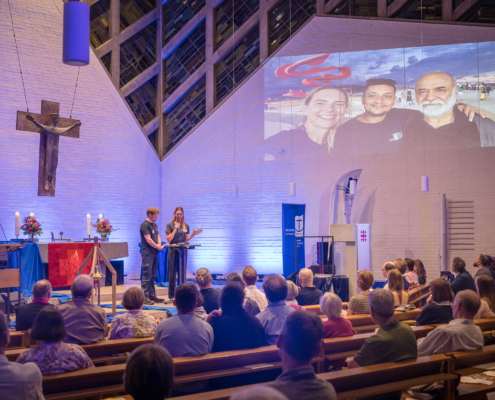 Das Foto zeigt den Innenraum der Auferstehungskirche in Hannover. Im Altarraum stehen Diakon Philipp Manthey und Kathrin Grund von der Seemannsmission in Bremerhaven und Santos. Kathrin Grund spricht gerade ins Mikrofon und stellt den Zuhörern ihre Arbeit in Brasilien vor. Im rechten oberen Rand des Bildes sieht an ein Foto von Kathrin Grund und zwei Kollegen am Strand von Santos. Es ist eine Abendveranstaltung, das Licht ist gedämpft und leicht bläulich.