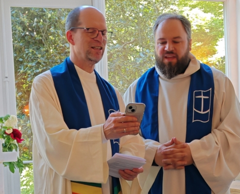 André Schuller (r.) und Matthias Ristau stehen beim Einführungsgottesdienst in weißen liturgischen Gewändern vorn und singen, während sie auf ein Smartphone schauen