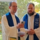 André Schuller (r.) und Matthias Ristau stehen beim Einführungsgottesdienst in weißen liturgischen Gewändern vorn und singen, während sie auf ein Smartphone schauen