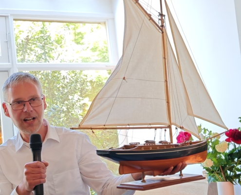 Pastor Stefan Leonhardt von der deutschen evangelischen Gemeinde Rotterdam steht mit einem Mikrofon in der einen und dem Modell vom Segelschiff in der anderen Hand vor einem Fenster zum Garten