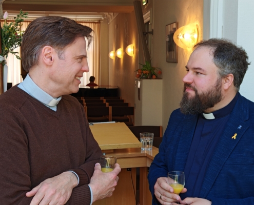 Die bieden Männer sind von vorn schräg zu sehen, wie sie im Gespräch einander gegenüber stehen, beide mir einem Glas Orangensaft in der Hand