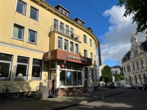 Das Bremerhavener Seemannshotel Portside Das Foto zeigt die Außenansicht des Seemannshotels Portside in Bremerhaven. Mittig kann man den Eingang erkennen, links führt eine kleine Treppe ins Innere. Die Fassade ist gelb gestrichen, im Kontrast dazu steht ein brauner Anstrich für den Foyerbereich. Die Sonne scheint, der Himmel ist blau, vereinzelt sind kleine Wölckchen zu sehen. Bäume werfen ihre Schatten auf den Eingang des Hotels.