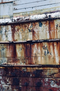 Das Foto zeigt eine Nahaufnahme eines Schiffswracks. Die ehemals weiße Farbe der Bordwand blättert an vielen Stellen ab, ist durch Rost braun verfärbt und verlaufen.
