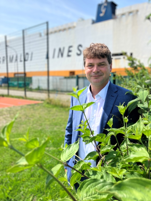 Zu sehen ist Prof. Dr. Iven Krämer, der auf dem Basketball-Feld des Seemannsclubs Welcome in Bremerhaven steht. Im Hintergrund fährt ein Containerschiff entlang. Es ist Frühjahr, man sieht das frische Grün eines Busches. Iven Krämer lächelt in die Kamera, die Sonne scheint.