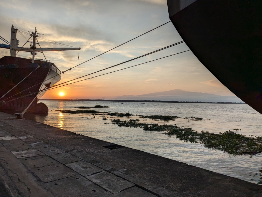 Auf dem Foto sieht man einen Sonnenuntergang im Hafen von Duoala in Kamerun. Gegen die Sonne sieht man dunkler zwei Schiffsrümpfe. Beide Schiffe sind mit Tauen an der Kaikante festgemacht. Auf dem Wasser treiben Algen, die Oberfläche ist sehr ruhig. Im Bildhintergrund sieht man Mount Cameroon. Die Atmosphäre auf dem Foto ist sehr stimmungsvoll und friedlich.