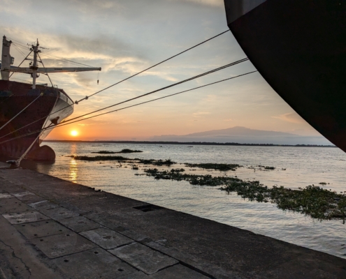 Auf dem Foto sieht man einen Sonnenuntergang im Hafen von Duoala in Kamerun. Gegen die Sonne sieht man dunkler zwei Schiffsrümpfe. Beide Schiffe sind mit Tauen an der Kaikante festgemacht. Auf dem Wasser treiben Algen, die Oberfläche ist sehr ruhig. Im Bildhintergrund sieht man Mount Cameroon. Die Atmosphäre auf dem Foto ist sehr stimmungsvoll und friedlich.
