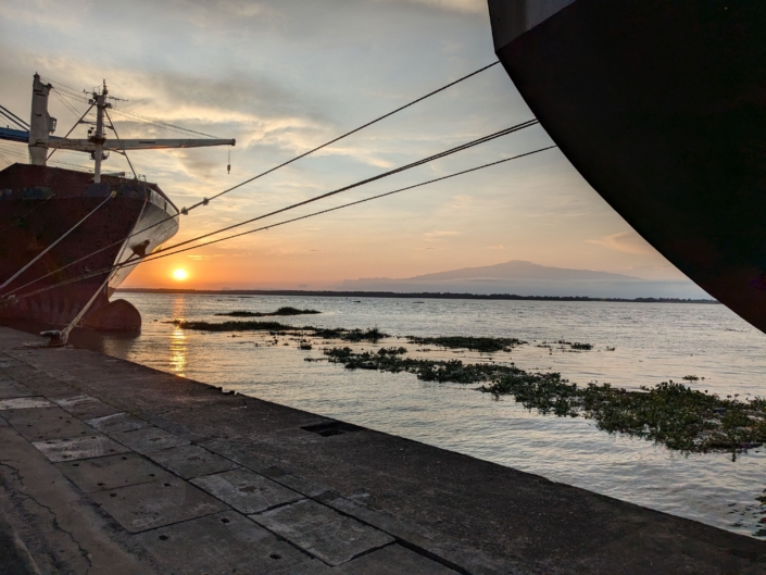 Auf dem Foto sieht man einen Sonnenuntergang im Hafen von Duoala in Kamerun. Gegen die Sonne sieht man dunkler zwei Schiffsrümpfe. Beide Schiffe sind mit Tauen an der Kaikante festgemacht. Auf dem Wasser treiben Algen, die Oberfläche ist sehr ruhig. Im Bildhintergrund sieht man Mount Cameroon. Die Atmosphäre auf dem Foto ist sehr stimmungsvoll und friedlich.