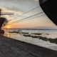 Auf dem Foto sieht man einen Sonnenuntergang im Hafen von Duoala in Kamerun. Gegen die Sonne sieht man dunkler zwei Schiffsrümpfe. Beide Schiffe sind mit Tauen an der Kaikante festgemacht. Auf dem Wasser treiben Algen, die Oberfläche ist sehr ruhig. Im Bildhintergrund sieht man Mount Cameroon. Die Atmosphäre auf dem Foto ist sehr stimmungsvoll und friedlich.