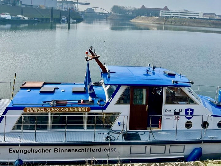 Das Bild zeigt das Kirchenboot Hinrich Wichern der Evangelischen Binnenschifferseelsorge/Deutschen Seemannsmission Duisburg im Duisburger Hafen. Das Dach des Bootes ist in einem hellen Blau lackiert, der untere Teil in Weiß, mit dem blauen Logo der Deutschen Seemannsmission.