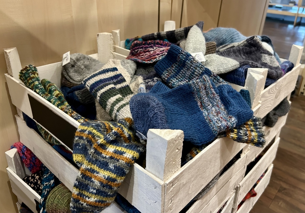 Zu sehen ist eine weiß gestrichene Holzkiste mit zahlreichen handgestrickten Socken in verschiedenen Mustern. Die meisten Socken sind in dunkleren Blau- und Scharztönen gehalten, manche sind aber auch von Weißtönen unterbrochen. Seeleute können sich die gespendeten Socken im Seemannsclub mitnehmen, um sich an Bord warmzuhalten.