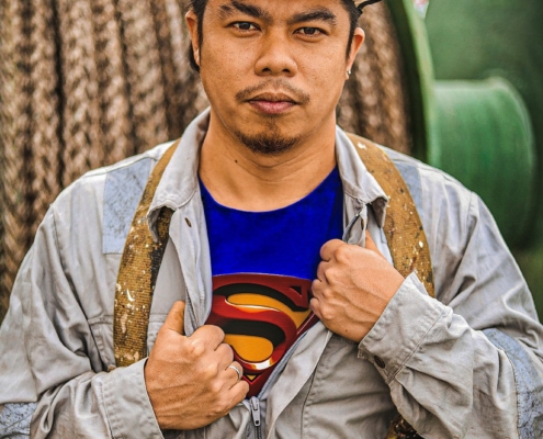 Das Foto zeigt einen jungen philippinischen Seemann, der ernst in die Kamera blickt. Auffällig ist sein blaues T-Shirt, das unter dem vermutlich ölverschmierten grauen Arbeitsoverall herausschaut, weil der Reußverschluss etwas geöffnet wurde. Es ist ein Superman-T-Shirt, das zu sehen ist. Der Fotograf heißt Aljon Manlangit, das Foto hat er "Ich bin Seemann und ich bin ein Held" genannt. Im Hintergrund sieht man eine grüne Trommel, wahrscheinlich mit einem aufgerolltem Tau oder einer dicken Kette. Der Seemann trägt einen weißen Schutzhelm auf dem Kopf. Auf seinen Schultern liegt locker ein mit weißer Farbe verschmierter Gurt, an dessen Ende Metallösen angebracht sind.