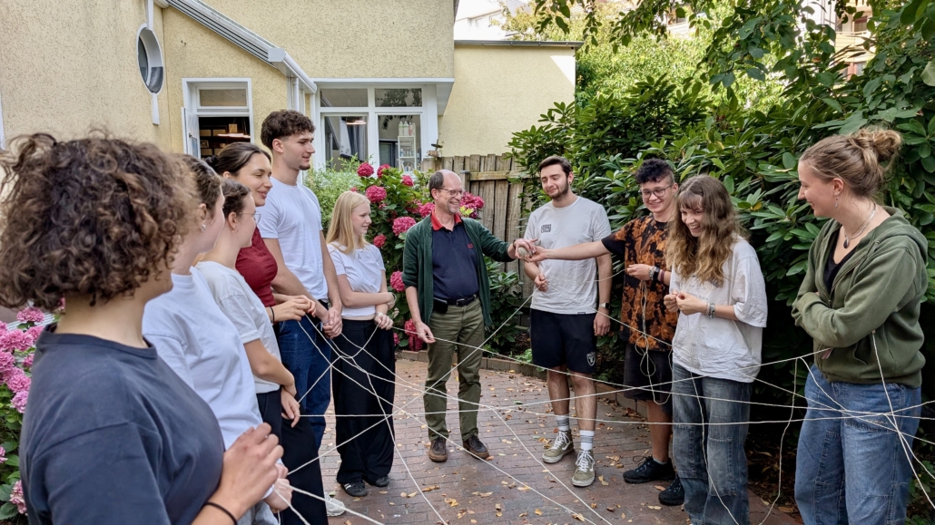 Das Foto zeigt eine Gruppe junger Leute, in deren Mitte Generalsekretär Matthias Ristau. Die Gruppe steht im Garten des Seemannshotels Portside in Bremerhaven. Es sind Teilnehmer des Seminars für Auslandsfreiwillige. Es ist Juli. Alle haben Enden von Fäden in ihren Händen, die kreuz und quer vernetzt und verknotet sind. Ziel des Spiels ist die Zusammenarbeit im Team. Das Spiel scheint der Gruppe Spaß zu machen.