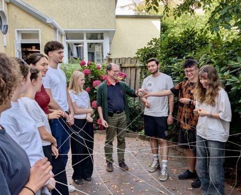 Das Foto zeigt eine Gruppe junger Leute, in deren Mitte Generalsekretär Matthias Ristau. Die Gruppe steht im Garten des Seemannshotels Portside in Bremerhaven. Es sind Teilnehmer des Seminars für Auslandsfreiwillige. Es ist Juli. Alle haben Enden von Fäden in ihren Händen, die kreuz und quer vernetzt und verknotet sind. Ziel des Spiels ist die Zusammenarbeit im Team. Das Spiel scheint der Gruppe Spaß zu machen.