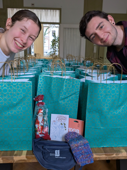 Alma Bloech und Joel Dressler (re.) haben Weihnachtstüten für Seeleute im Amsterdamer Hafen gepackt. Sie lächeln freundlich in die Kamera und haben sich über viele Tüten gebeugt, die auf einem Tisch aufgereiht stehen.