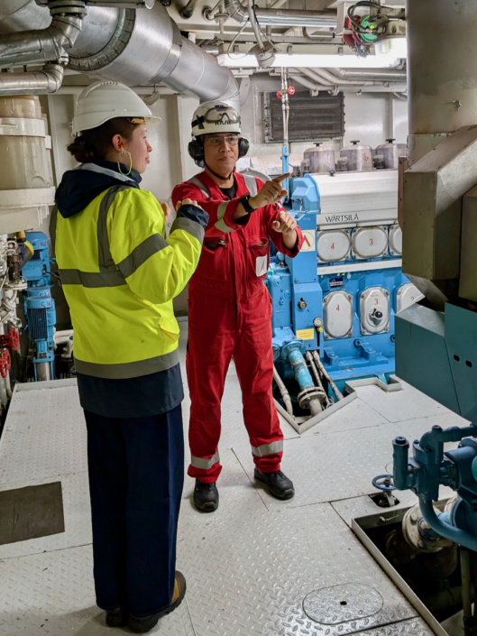 Alma Bloech (links) bei ihrem ersten Bordbesuch im Maschinenraum eines Schiffes im Hafen von Amsterdam. Rechts ist ein Seemann im roten Arbeitsoverall zu sehen. Die junge Freiwillige und der Seemann tragen beide weiße Schutzhelme und Gehörschutz.