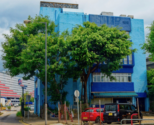 Das Foto zeigt das sogenannte Blue House im Hafen von Hongkong. Es beherbergt den ökumenischen Seemannsclub sowie Büros der verschiedenen Organisationen, die sich zum Wohl von Seeleuten in Hongkong engagieren. Das Gebäude ist in einem helleren und einem dunkleren Blauton gestrichen. Zwei Bäume säumen den Eingang des Gebäudes. Davor befindet sich ein Parkplatz.