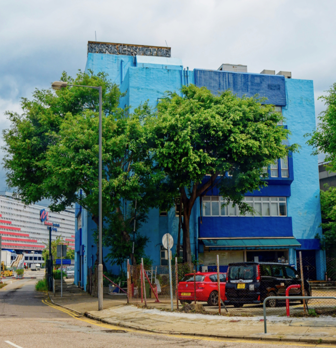 Das Foto zeigt das sogenannte Blue House im Hafen von Hongkong. Es beherbergt den ökumenischen Seemannsclub sowie Büros der verschiedenen Organisationen, die sich zum Wohl von Seeleuten in Hongkong engagieren. Das Gebäude ist in einem helleren und einem dunkleren Blauton gestrichen. Zwei Bäume säumen den Eingang des Gebäudes. Davor befindet sich ein Parkplatz.