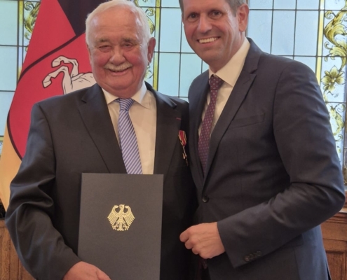 Ehrung für Wilfrid Adam, der links auf dem Bild steht, neben dem Ministerpräsidentin Olaf Lies. Beide tragen ein dunkles Sakko, Adam hält eine schwarze Mappe mit einem goldenen Bundesadler in den Händen
