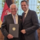 Ehrung für Wilfrid Adam, der links auf dem Bild steht, neben dem Ministerpräsidentin Olaf Lies. Beide tragen ein dunkles Sakko, Adam hält eine schwarze Mappe mit einem goldenen Bundesadler in den Händen