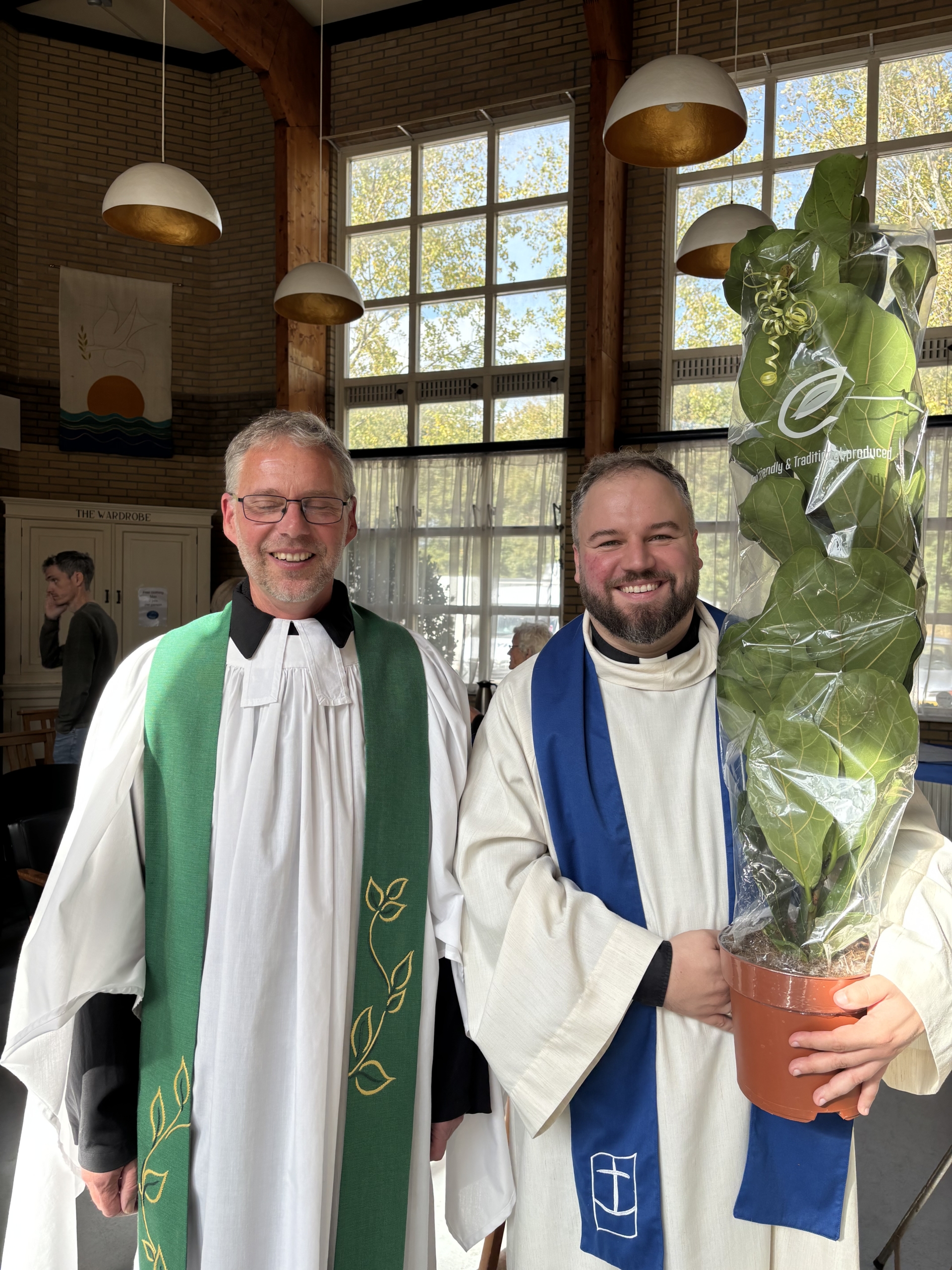 Links steht Pastor Stefan Leomhardt in Talar mit weißer Albe darüber udn grüner Stola, neben ihm André Schuller in weißer Albe mit blauer Stola mit dem Logo der Seemannsmission und einem Topf mit einer großen grünen Pflanze in der Hnd