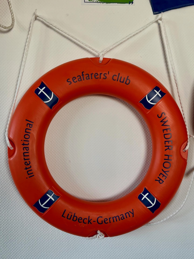 Das Foto zeigt einen orangenen Rettungsring mit der blauen Aufschrift International Seafarers' club Sweder Hoyer der Lübecker Seemannsmission vor einem weißen Hintergrund.