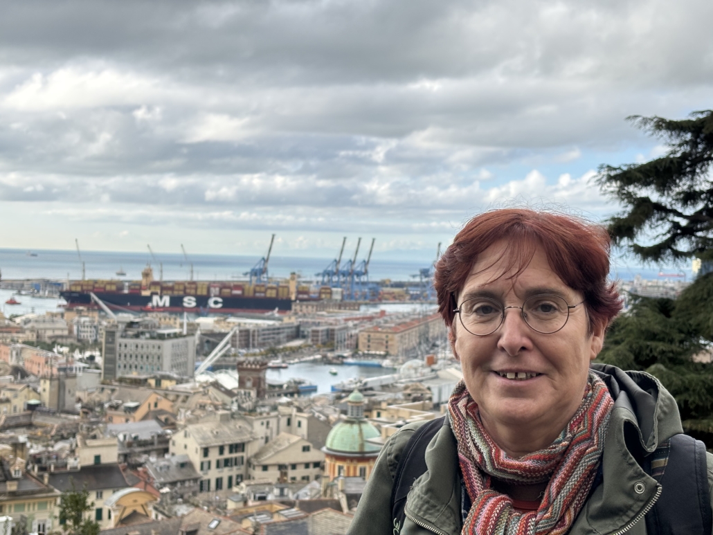Zu sehen ist Barbara Panzlau, die seit mehr als 20 Jahren im Hafen von Genua als Diakonin für Seeleute unterwegs ist. Barbara Panzlau hat rot gefärbtes Haar, das sie kurz trägt. Eine runde Brille sitzt auf ihrer Nase. Sie lächelt verhalten in die Kamera. Im Hintergrund ist der Hafen von Genua zu sehen. Dicke graue Wolken hängen am Himmel.