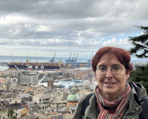 Zu sehen ist Barbara Panzlau, die seit mehr als 20 Jahren im Hafen von Genua als Diakonin für Seeleute unterwegs ist. Barbara Panzlau hat rot gefärbtes Haar, das sie kurz trägt. Eine runde Brille sitzt auf ihrer Nase. Sie lächelt verhalten in die Kamera. Im Hintergrund ist der Hafen von Genua zu sehen. Dicke graue Wolken hängen am Himmel.