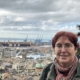 Zu sehen ist Barbara Panzlau, die seit mehr als 20 Jahren im Hafen von Genua als Diakonin für Seeleute unterwegs ist. Barbara Panzlau hat rot gefärbtes Haar, das sie kurz trägt. Eine runde Brille sitzt auf ihrer Nase. Sie lächelt verhalten in die Kamera. Im Hintergrund ist der Hafen von Genua zu sehen. Dicke graue Wolken hängen am Himmel.
