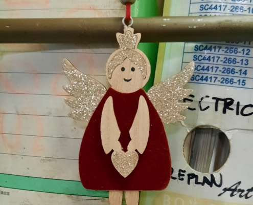 Das Foto zeigt einen Adventsengel an einer roten Schnur. Er hat goldverzierte Flügel, goldene Haare und trägt ein rotes Kleid. Die Figur lächelt. Sie hängt an Bord eines Schiffes vor Stehordnern mit Daten, die wahrscheinlich auf der Brücke oder in einem Büro wichtig sind.