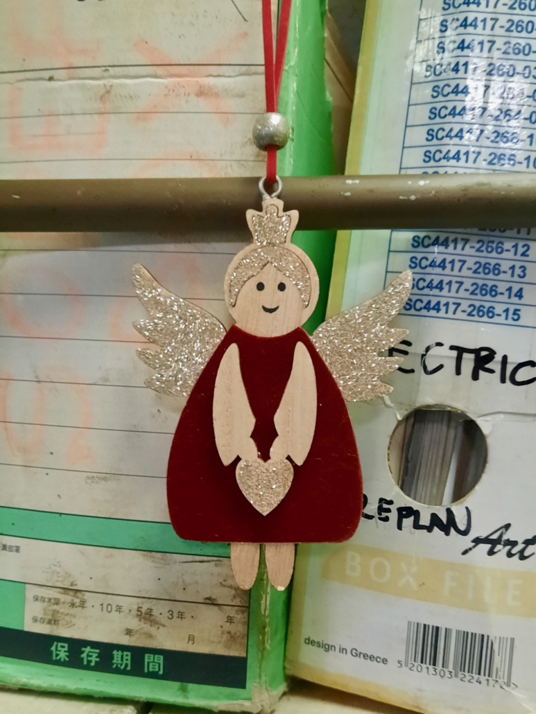 Das Foto zeigt einen Adventsengel an einer roten Schnur. Er hat goldverzierte Flügel, goldene Haare und trägt ein rotes Kleid. Die Figur lächelt. Sie hängt an Bord eines Schiffes vor Stehordnern mit Daten, die wahrscheinlich auf der Brücke oder in einem Büro wichtig sind.