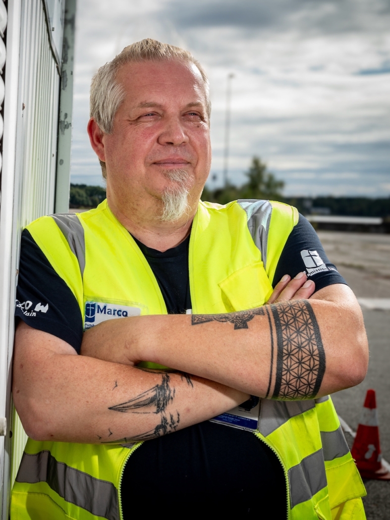 Das Foto zeigt Diakon Marco Folchnandt aus Lübeck im Lübecker Hafen, wie er an einer weißen Wand lehnt, die Arme vpr dem Körper verschränkt. Man kann gut seine Unterarm-Tattoos erkennen, auf dem rechten Unterarm ein Segelboot. Er trägt eine neongelbe Schutzweste und ein kurzärmeliges T-Shirt der Detuschen Seemannsmission.
