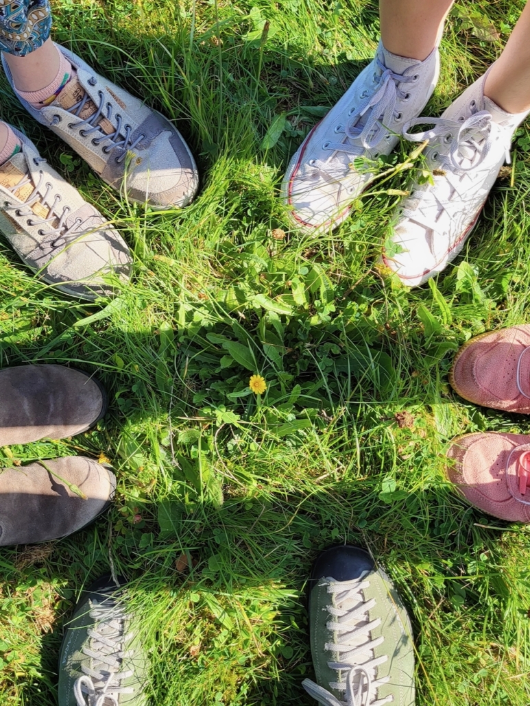 Das Foto zeigt den Blick vom Betrachter auf eine grüne Sommerwiese. Zu sehen sind nur die Schuhe von fünf Personen, mindestens drei davon sind junge Menschen, was man an den Schuhen erkennen könnte. Die Sonne scheint und in der Mitte des Kreises, den die fünf Menschen bilden, wächst eine kleine gelbe Blume auf der Wiese.