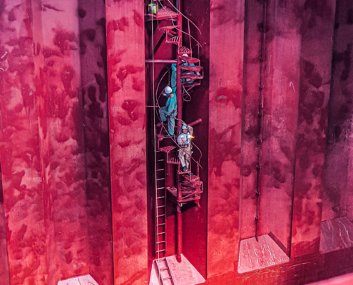 Im gezeigten Foto an einer Bordwand eines Schiffes dominiert die Farbe Magenta. Von rechts oeben changieren die Farben von Lila in einen Rotton links unten. Man sieht drei Seeleute in Arbeitsoveralls, die eine Wendeltreppe erklimmen. Ein Farbeimer steht auf der obersten Stufe der Treppe. Die drei Männer tragen Schutzhelme. Wahrscheinlich werden sie an der Bordwand streichen oder haben das bereits getan.