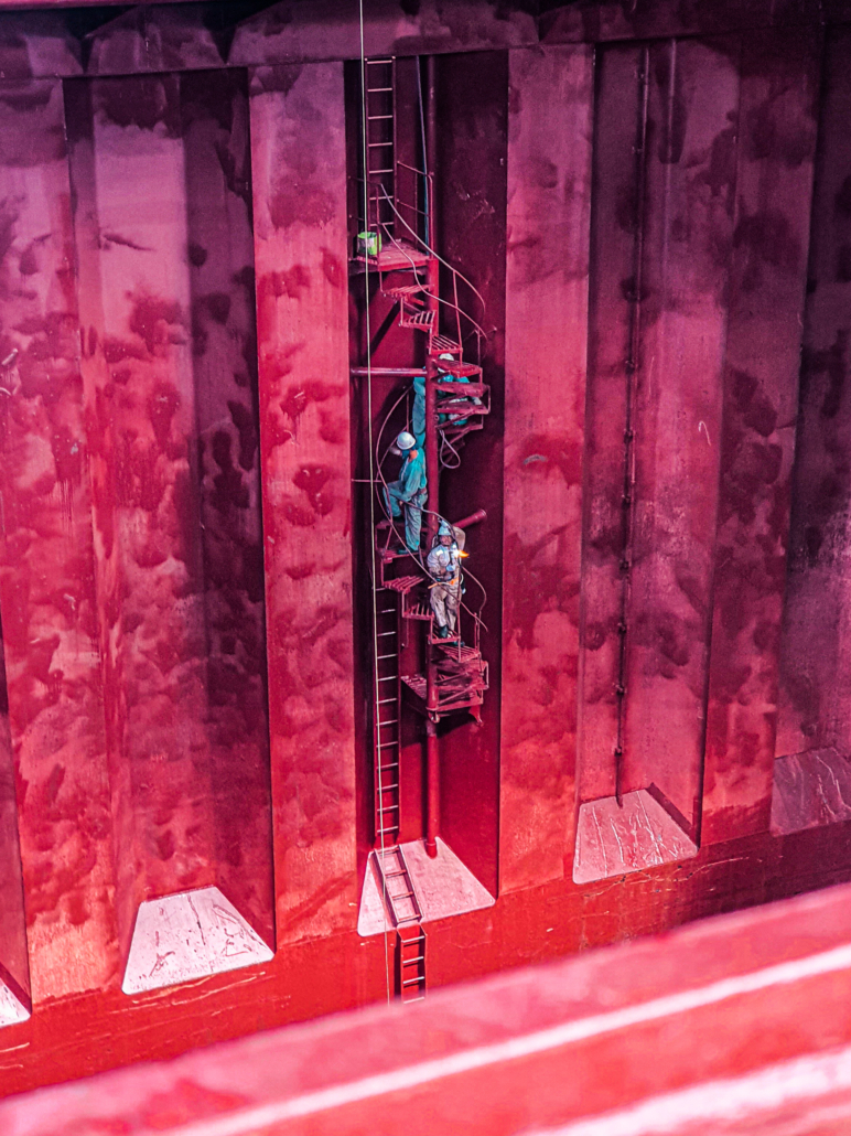 Im gezeigten Foto an einer Bordwand eines Schiffes dominiert die Farbe Magenta. Von rechts oeben changieren die Farben von Lila in einen Rotton links unten. Man sieht drei Seeleute in Arbeitsoveralls, die eine Wendeltreppe erklimmen. Ein Farbeimer steht auf der obersten Stufe der Treppe. Die drei Männer tragen Schutzhelme. Wahrscheinlich werden sie an der Bordwand streichen oder haben das bereits getan.