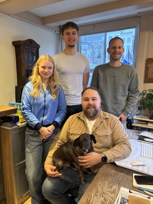 Das Foto zeigt das Team der Deutschen Seemannsmission Amsterdam. Die vier Mitglieder waren zur Aufnahme des Fotos gemeinsam im Büro des alten Grachtenhauses Keizersgracht 733. Im Vordergrund sitzt Diakon Thomas Kirschner. Auf seinem Schoß sitzt sein Hund Lotti. Die beiden internationalen Freiwilligen sind links und mittig in der zweiten Reihe zu sehen. Alle vier lächeln in die Kamera.