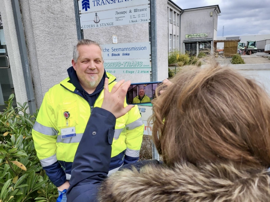 Marco Folchnandt, Diakon der Deutschen Seemannsmission Lübeck, wird im Lübecker Hafen vor dem Eingang zum Bürokomplex fotografiert. Er lächelt freundlich in die Handykamera. Der Fotograf steht hinter der Frau, die gerade ein Foto des Diakons macht.