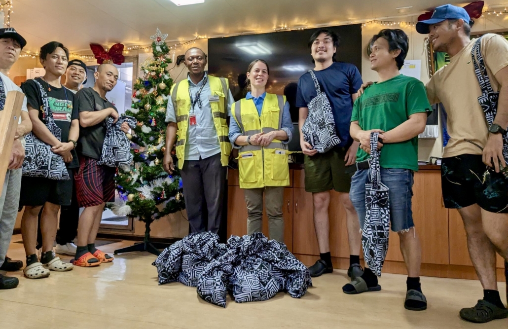 Das Foto zeigt einen Besuch an Bord eines Schiffes in der Vorweihnachtszeit. Die Crew-Messe an Bord ist weihnachtliche geschmückt. Die Seemänner stehen rings um die Bordbesucher der Deutschen Seemannsmission Douala. Zu Füßen von Diakonin Ina Bammann liegen dunkelblaue, genähte Weihnachtsbeutel mit traditionellen afrikanischen Mustern in weiß gehalten. Die Seeleute werden mit Weihnachtsliedern und den Tüten beschenkt, weil viele von ihnen Weihnachten auf See verbringen werden.