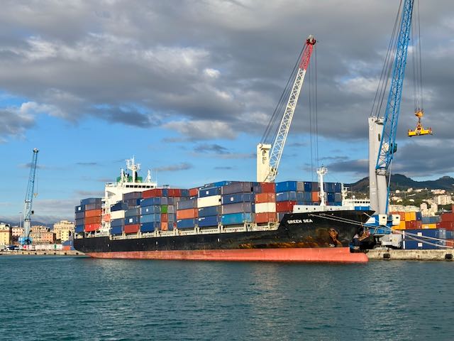 Das Foto zeigt den Hafen von Genua und ein Containerschiff mit vielen Containern in blau, orange, weiß und rot, das gerade be- oder entladen wird. Direkt über dem Schiff befindet sich eine gewaltige graue Wolke, obwohl das Foto an einem sonst sonnigen Tag aufgenommen wurde.