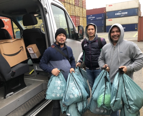 Das Foto zeigt drei phlilippinische Seeleute, die vor einem kleinen Shuttelbus stehen, dessen Tür geöffnet ist. Im Inneren kann man Gepäck, wie z. B. Kartons wie in einem Lieferwagen sehen. Die drei jungen Männer halten prall gefüllte, fast durchsichtige hellblaue Kunststofftüten in den Händen. Das sind die Weihnachtstüten der Deutschen Seemannsmission New York. Im Hintergrund sind an Land gestapelte Container in gelb, rot, blau, weiß zu sehen. Bis zu vier Container sind übereinnander gestapelt.