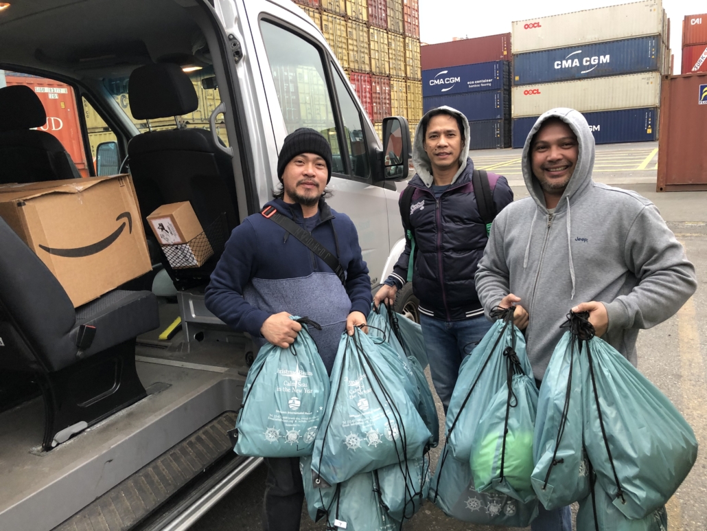 Das Foto zeigt drei philippinische Seeleute, zwei haben eine Kapuze auf, der Mann ganz links eine Wollmütze. Die drei jungen Männer halten Weihnachtstüten der Deutschen Seemannsmission New York in den Händen, die sie im Hafen von Newark mit dem Shuttlebus an Bord ihres Schiffes nehmen werden. Im Hintergrund sieht man gestapelte Container in allerlei Farben. Die Tür des kleinen Shuttlebusses steht auf, man sieht Gepäck im Inneren des Wagens.