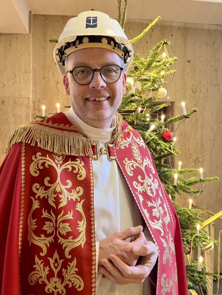 Diakon Henry Schwier, im Gewand des Heiligen Nikolaus, mit einem weißen Schutzhelm der Deutschen Seemannsmission vor einem festlich geschmückten Weihnachtsbaum.