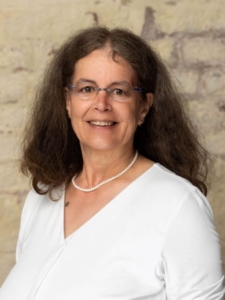 Das Foto ist ein Portrait von Diakonin Martina Platte, die mit offenen, langen, lockigen dunkelbraunen Haaren und weißem Oberteil freundlich in die Kamera lächelt. Sie trägt eine weiße Perlenkette, der Anlass ist festlich.