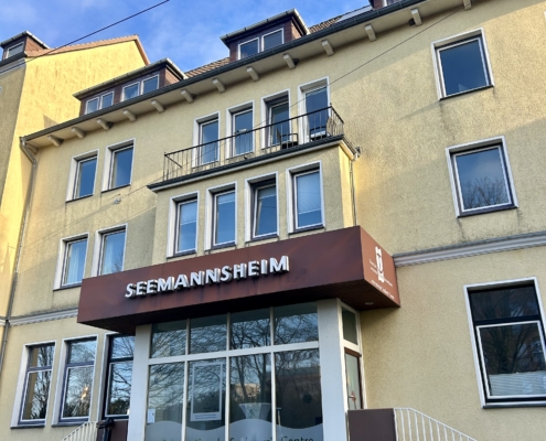 Das Foto zeigt das Seemannshotel Portside in der Schifferstraße 51 in Bremerhaven. Das Haus mit Übernachtungsmöglichkeiten und Aufenthaltsräumen für Seeleute wird nach 125 Jahren zum Ende des Jahres 2025 geschlossen. Die Fassage erstrahlt in einem freundlichen Gelb. Über dem Eingang prangt der Schriftzug "Seemannsheim" in großen weißen Lettern.