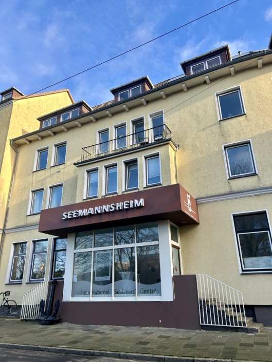 Das Foto zeigt das Seemannshotel Portside in der Schifferstraße 51 in Bremerhaven. Das Haus mit Übernachtungsmöglichkeiten und Aufenthaltsräumen für Seeleute wird nach 125 Jahren zum Ende des Jahres 2025 geschlossen. Die Fassage erstrahlt in einem freundlichen Gelb. Über dem Eingang prangt der Schriftzug "Seemannsheim" in großen weißen Lettern.