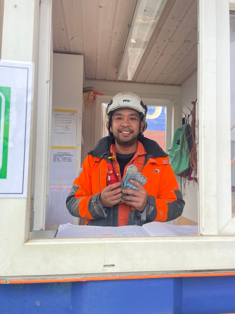 Das Foto zeigt einen philippinischen Seemann in Arbeitsoverall mit weißem Schutzhelm auf dem Kopf. Der junge Mann hölt einen Schokoladen-Weihnachtsmann und ein paar selbstgestrickte Socken in den Händen. Er freut sich und lächelt in die Kamera.